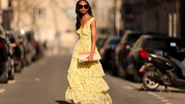 Emilie Joseph @in_fashionwetrust wears black sunglasses, a pale yellow with green and yellow flower print pattern V-neck / tank-top / backless / belted / ruffled maxi long dress, a beige fabric ruffled clutch, silver rings, pale yellow matte leather pointed pumps heels shoes, during a street style fashion photo session, on March 26, 2022 in Paris, France.