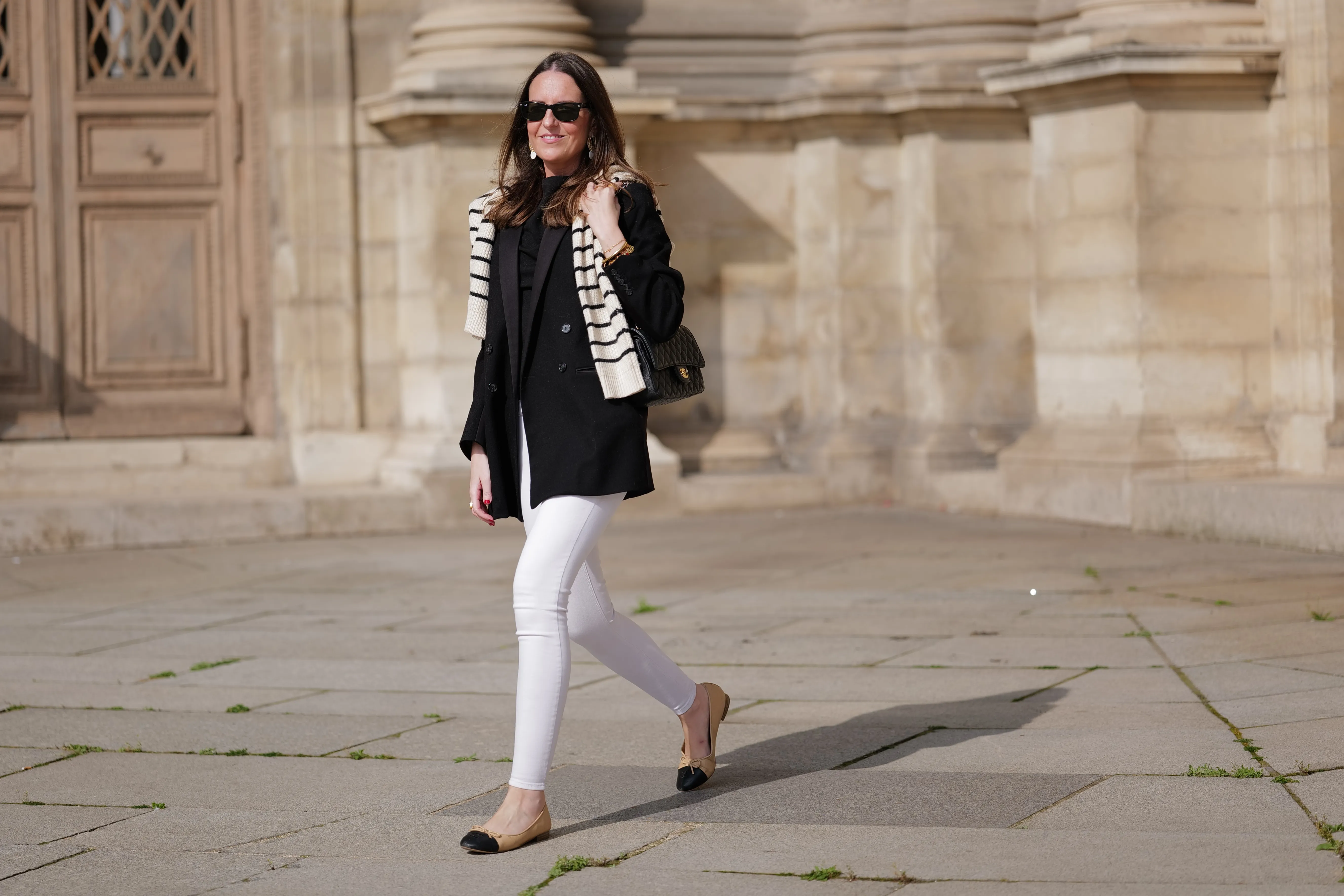 Alba Garavito Torre wears black sunglasses from Ray Ban, gold and white pendant large earrings, a black t-shirt, a white with black small striped print pattern wool pullover from Massimo Dutti, a black shiny grained leather Timeless shoulder bag from Chanel, a black oversized blazer jacket from Munthe, white skinny denim pants from Zara, beige shiny leather with black toe-cap ballerinas from Chanel , during a street style fashion photo session, on April 29, 2023 in Paris, France.