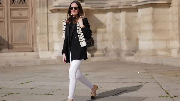 Alba Garavito Torre wears black sunglasses from Ray Ban, gold and white pendant large earrings, a black t-shirt, a white with black small striped print pattern wool pullover from Massimo Dutti, a black shiny grained leather Timeless shoulder bag from Chanel, a black oversized blazer jacket from Munthe, white skinny denim pants from Zara, beige shiny leather with black toe-cap ballerinas from Chanel , during a street style fashion photo session, on April 29, 2023 in Paris, France.