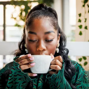 A young woman indulges in the aroma of her coffee, closing her eyes to savor the moment in a leafy cafe setting.