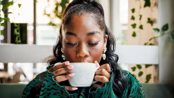 A young woman indulges in the aroma of her coffee, closing her eyes to savor the moment in a leafy cafe setting.