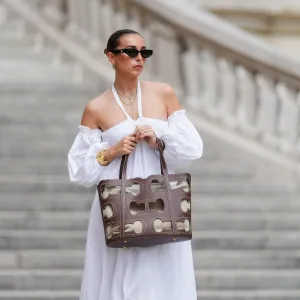 Gabriella Berdugo wears sunglasses, earrings, a white off-shoulder linen gathered dress from kalita with balloons / puff sleeves, a brown summer bag from Carolina Herrera in brown leather, during a street style fashion photo session, on May 12, 2024 in Paris, France.