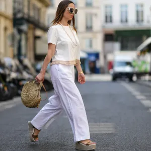PARIS, FRANCE - MAY 10: Maria Rosaria Rizzo wears white sunglasses, a white pearls long necklace, a white latte t-shirt with fringed borders, a beige wickers handbag from Guerlain, a gold large bracelet, gold rings, high waist white wide legs linen pants, gold shiny leather strappy with wicker wedge heels sandals, during a street style fashion photo session, on May 10, 2022 in Paris, France.