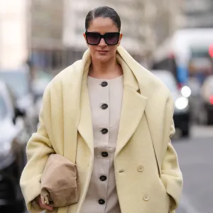 PARIS, FRANCE - MARCH 09: Gili Biegun wears black sunglasses, cream buttoned blazer jacket, light yellow oversized double breasted wool coat, dark yellow scarf, light brown suede bag, outside Akris, during the Paris Fashion week Women's Fall/Winter 2025-2026 on March 9, 2025 in Paris, France. (Photo by Edward Berthelot/Getty Images)