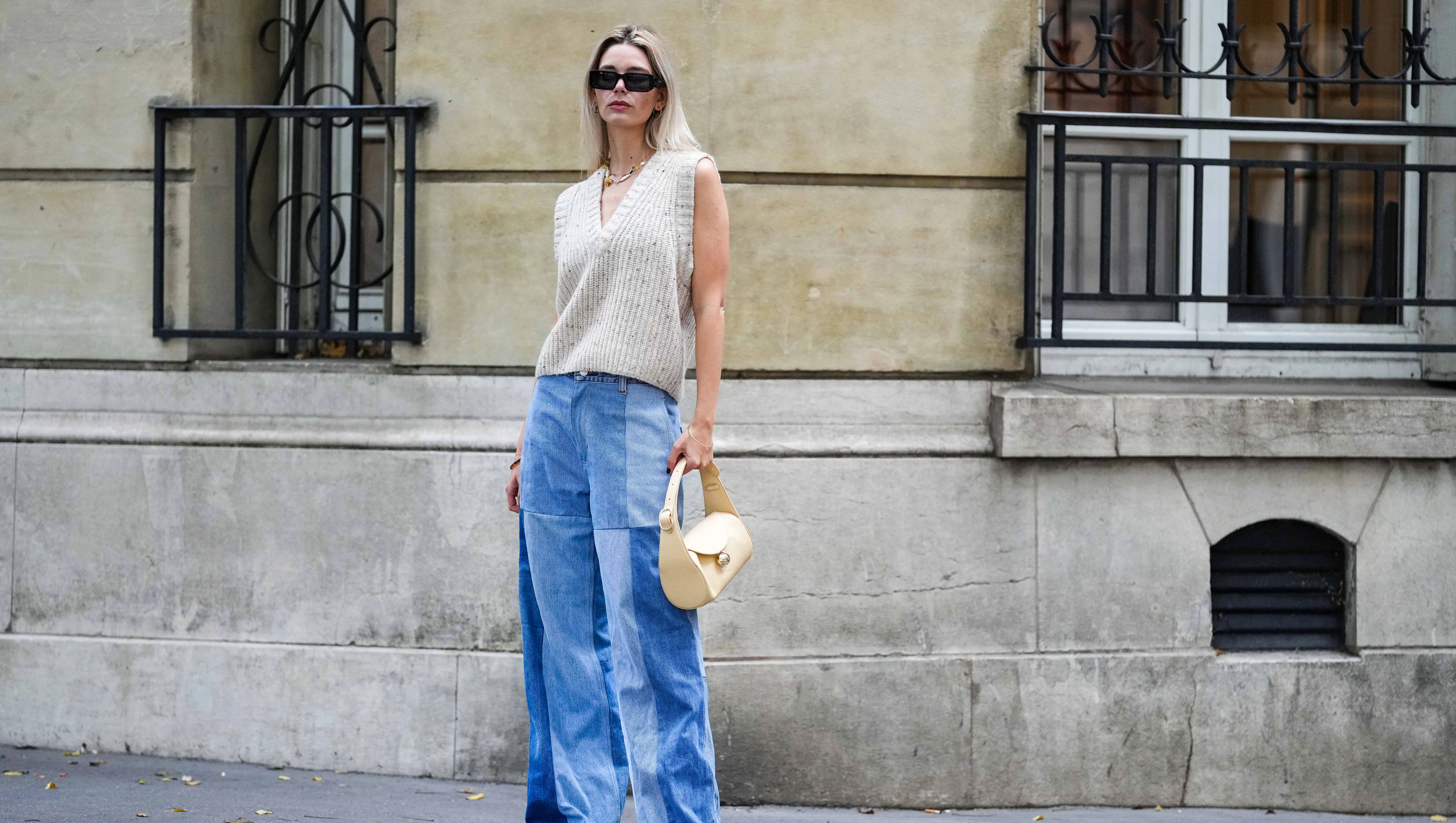 PARIS, FRANCE - OCTOBER 18: Natalia Verza wears black sunglasses, silver earrings, a white pearls / yellow necklace, a white latte wool V-neck / sleeveless pullover, a brown bracelet, a gold bracelet, a beige shiny leather handbag, pale blue bicolored yoke denim wide legs pants, black shiny leather pointed pumps heels shoes , during a street style fashion photo session, on October 18, 2022 in Paris, France.
