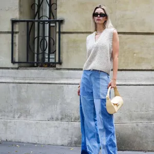 PARIS, FRANCE - OCTOBER 18: Natalia Verza wears black sunglasses, silver earrings, a white pearls / yellow necklace, a white latte wool V-neck / sleeveless pullover, a brown bracelet, a gold bracelet, a beige shiny leather handbag, pale blue bicolored yoke denim wide legs pants, black shiny leather pointed pumps heels shoes , during a street style fashion photo session, on October 18, 2022 in Paris, France.