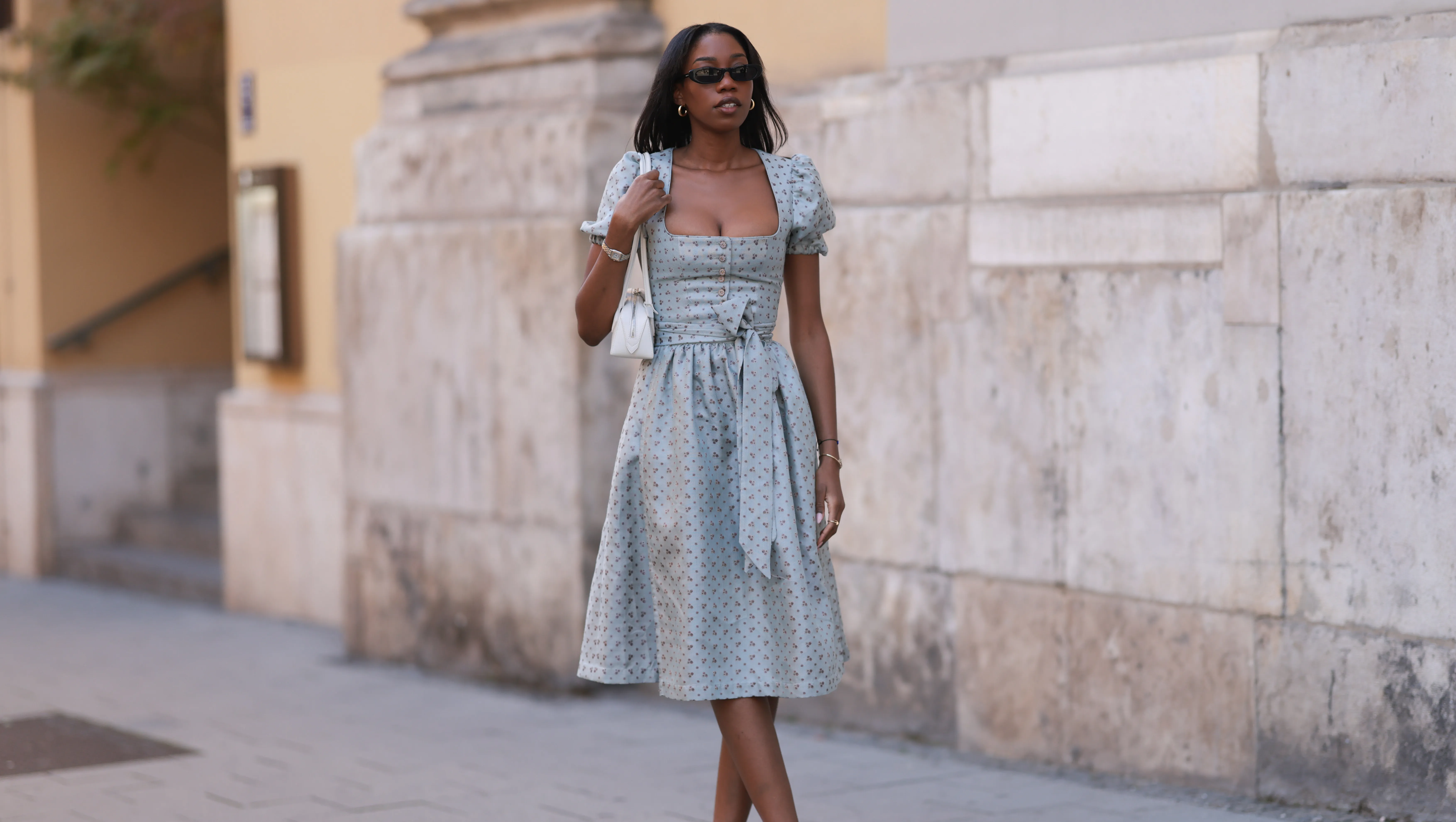 MUNICH, GERMANY - SEPTEMBER 21: Noutcha Lopes is seen wearing narrow rectangular sunglasses with a rounded black frame from Saint Laurent; chunky gold hoop earrings; a light blue dirndl with a flower pattern, a balconette neckline, a button placket, puffy short sleeves, and a bow from Limberry; a watch in silver and gold from Cartier; the yellow gold “Juste un Clou“ bracelet and ring from Cartier; a black bracelet with a blue eye pendant; a rectangular white leather “Le Teckle“ bag with tubular handles from Alaïa; white ankle socks; and pointed shiny black leather pumps during the opening day of the 2024 Munich Oktoberfest on September 21, 2024 in Munich, Germany. This year's Oktoberfest will run through October 6 and is expected to draw millions of visitors. (Photo by Jeremy Moeller/Getty Images)