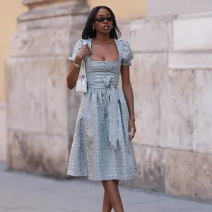 MUNICH, GERMANY - SEPTEMBER 21: Noutcha Lopes is seen wearing narrow rectangular sunglasses with a rounded black frame from Saint Laurent; chunky gold hoop earrings; a light blue dirndl with a flower pattern, a balconette neckline, a button placket, puffy short sleeves, and a bow from Limberry; a watch in silver and gold from Cartier; the yellow gold “Juste un Clou“ bracelet and ring from Cartier; a black bracelet with a blue eye pendant; a rectangular white leather “Le Teckle“ bag with tubular handles from Alaïa; white ankle socks; and pointed shiny black leather pumps during the opening day of the 2024 Munich Oktoberfest on September 21, 2024 in Munich, Germany. This year's Oktoberfest will run through October 6 and is expected to draw millions of visitors. (Photo by Jeremy Moeller/Getty Images)