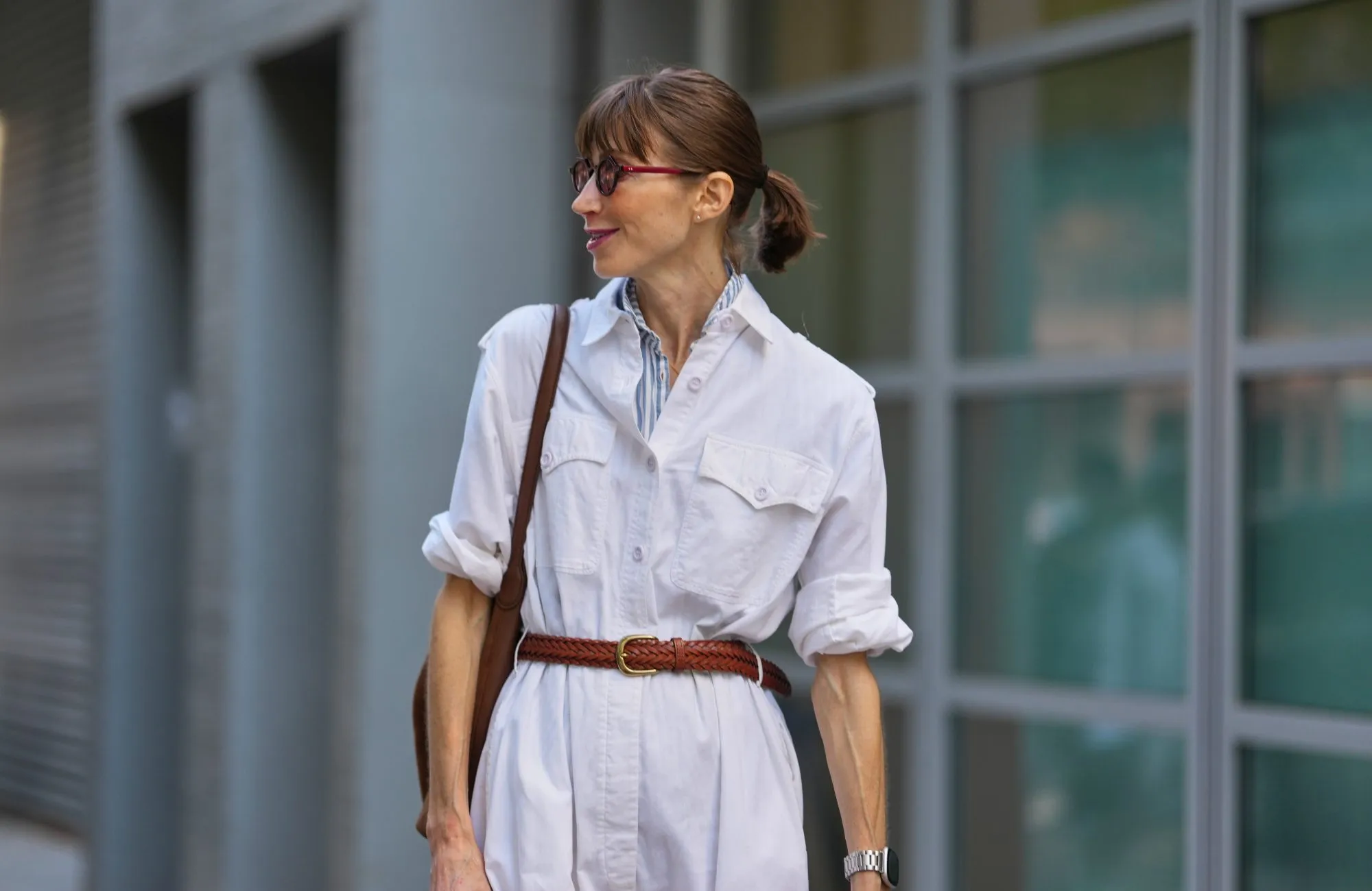NEW YORK, NEW YORK - SEPTEMBER 15: A guest wears their hair pulled back into a low ponytail with straight bangs, small round burgundy sunglasses with dark lenses, a silver metal watch with a black face on the left wrist, a brown leather shoulder bag with a thin strap, a white button-front jumpsuit in a lightweight woven fabric with chest flap pockets and rolled sleeves, a blue-and-white striped shirt collar peeking at the neckline, a braided brown leather belt with a gold-toned buckle cinching the waist, outside Zankov, during New York Fashion Week, on September 15, 2025 in New York, New York (Photo by Edward Berthelot/Getty Images)