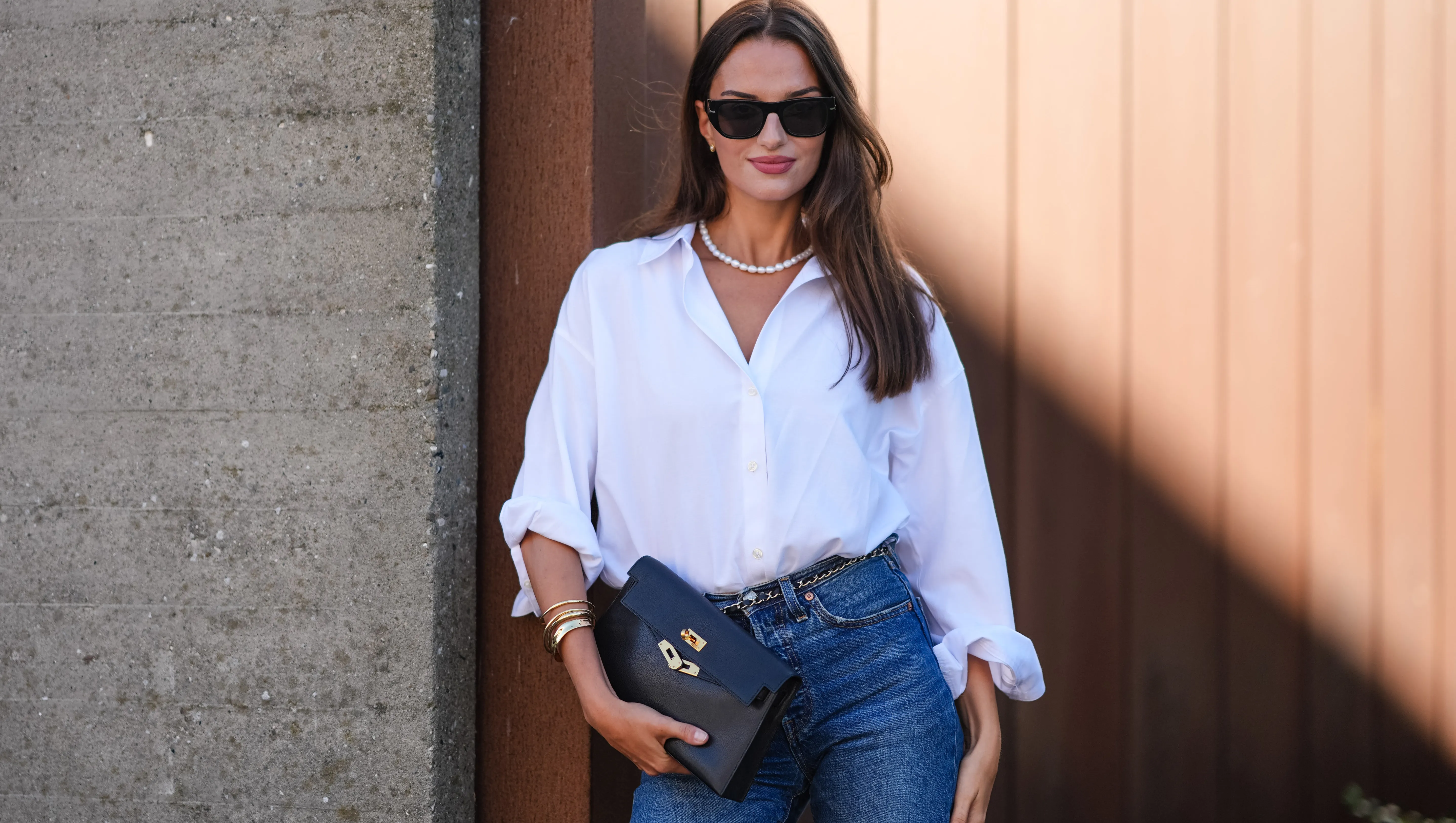 COPENHAGEN, DENMARK - AUGUST 6: Gabrielle Caunesil Pozzoli wears black sunglasses, white beaded necklace, white button up long sleeve rolled up shirt, black leather bag, gold and black belt, gold rings, light blue denim jean pants, outside Remain, during the Copenhagen Fashion Week Spring/Summer 2024-2025 on August 6, 2024 in Copenhagen, Denmark. (Photo by Edward Berthelot/Getty Images)