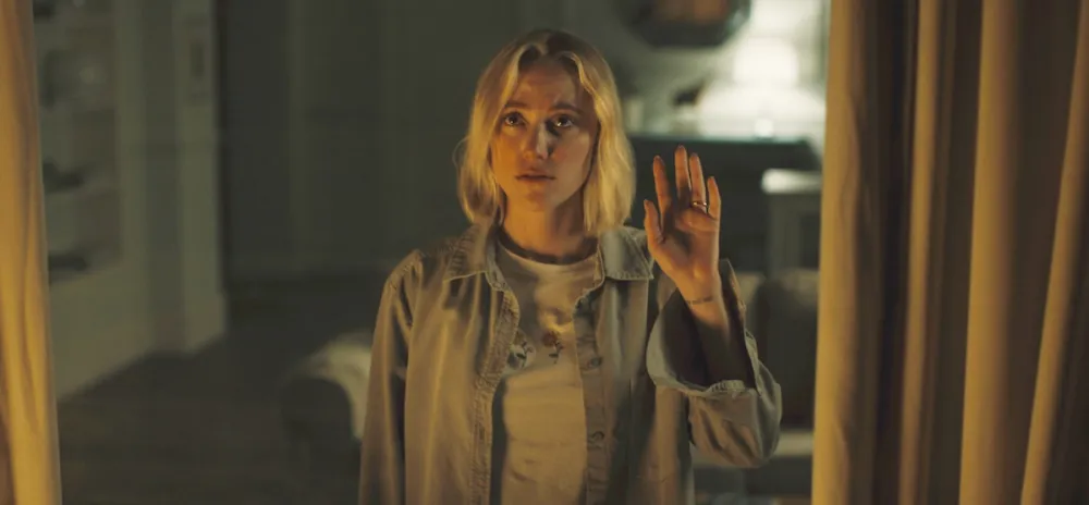 A woman waves while looking out a window in Watcher.