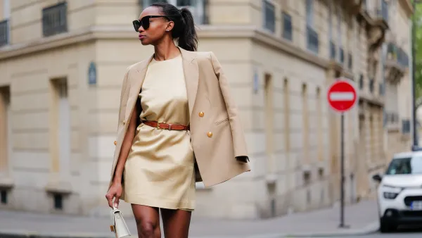 PARIS, FRANCE - APRIL 30: Emilie Joseph wears black large sunglasses, a pale yellow high neck / sleeveless / short dress, a camel shiny leather small belt, a beige with gold buttons blazer jacket, a beige wicker and white shiny leather bicolored handbag, pale yellow matte leather block heels / high knees boots , during a street style fashion photo session, on April 30, 2023 in Paris, France. (Photo by Edward Berthelot/Getty Images)