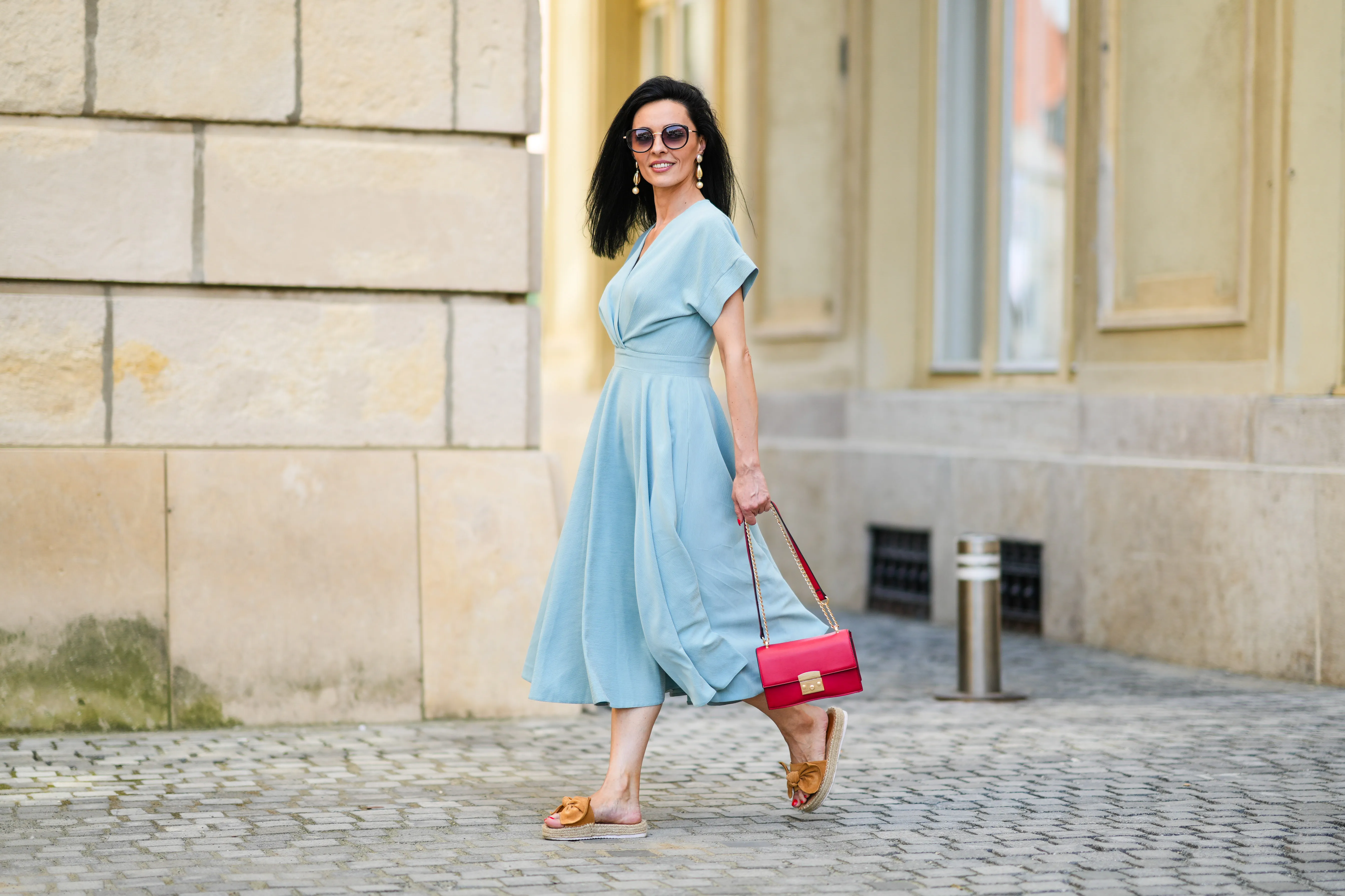 Cosmina Pasarin wears sunglasses, a pastel blue gathered low-neck midi dress, a red bag, brown sandals with platform beige wicker platforms, on July 27, 2021 in Sibiu, Romania.