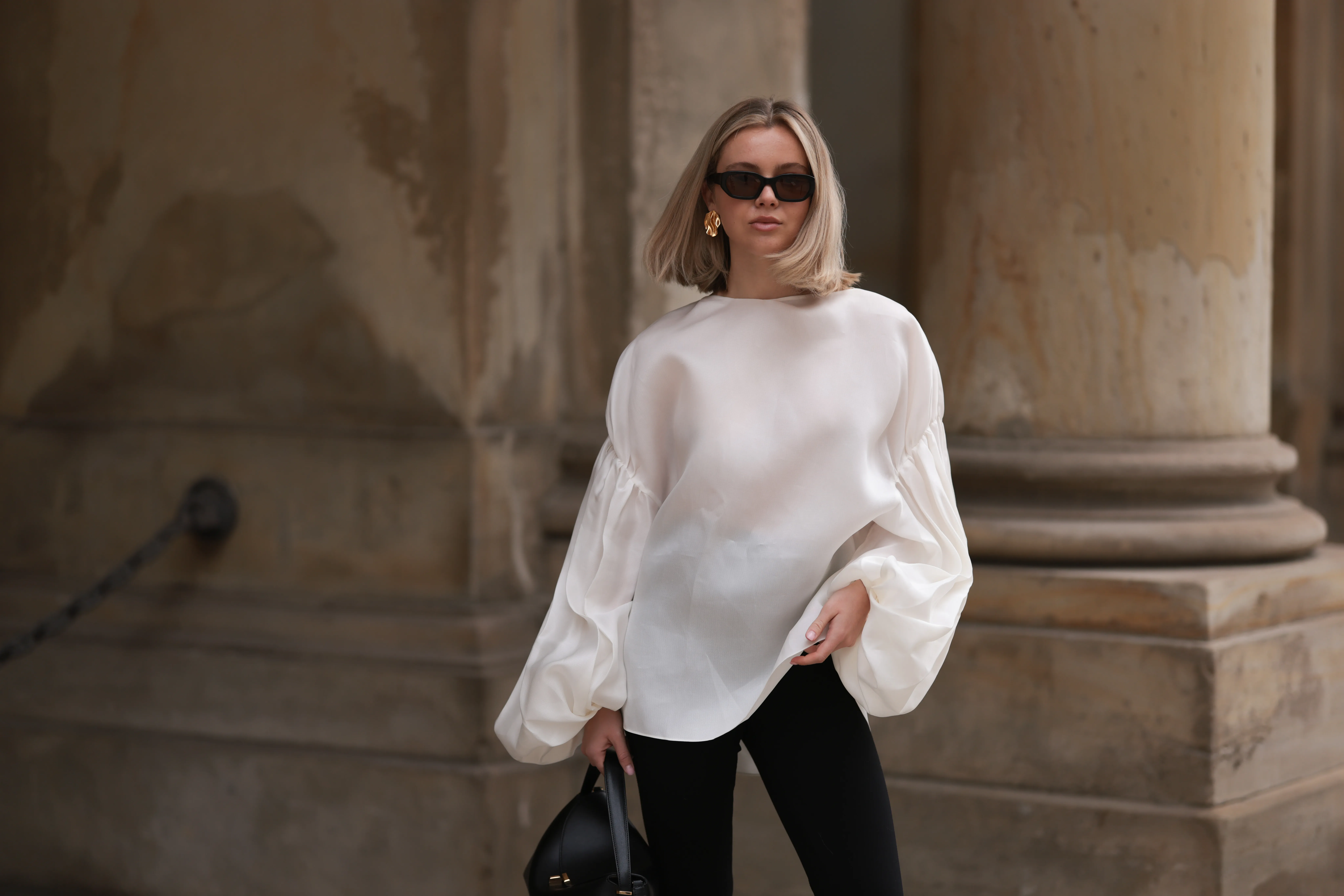 Isabelle Hartmann seen wearing Vehla Eyewear black / brown sunglasses, Nootka gold earrings, Khaite oversized puffy ruffled long sleeve silk top, Isabelle Hartmann x Edited black slit pants and Toteme black leather bag on April 05, 2024 in Hamburg, Germany.