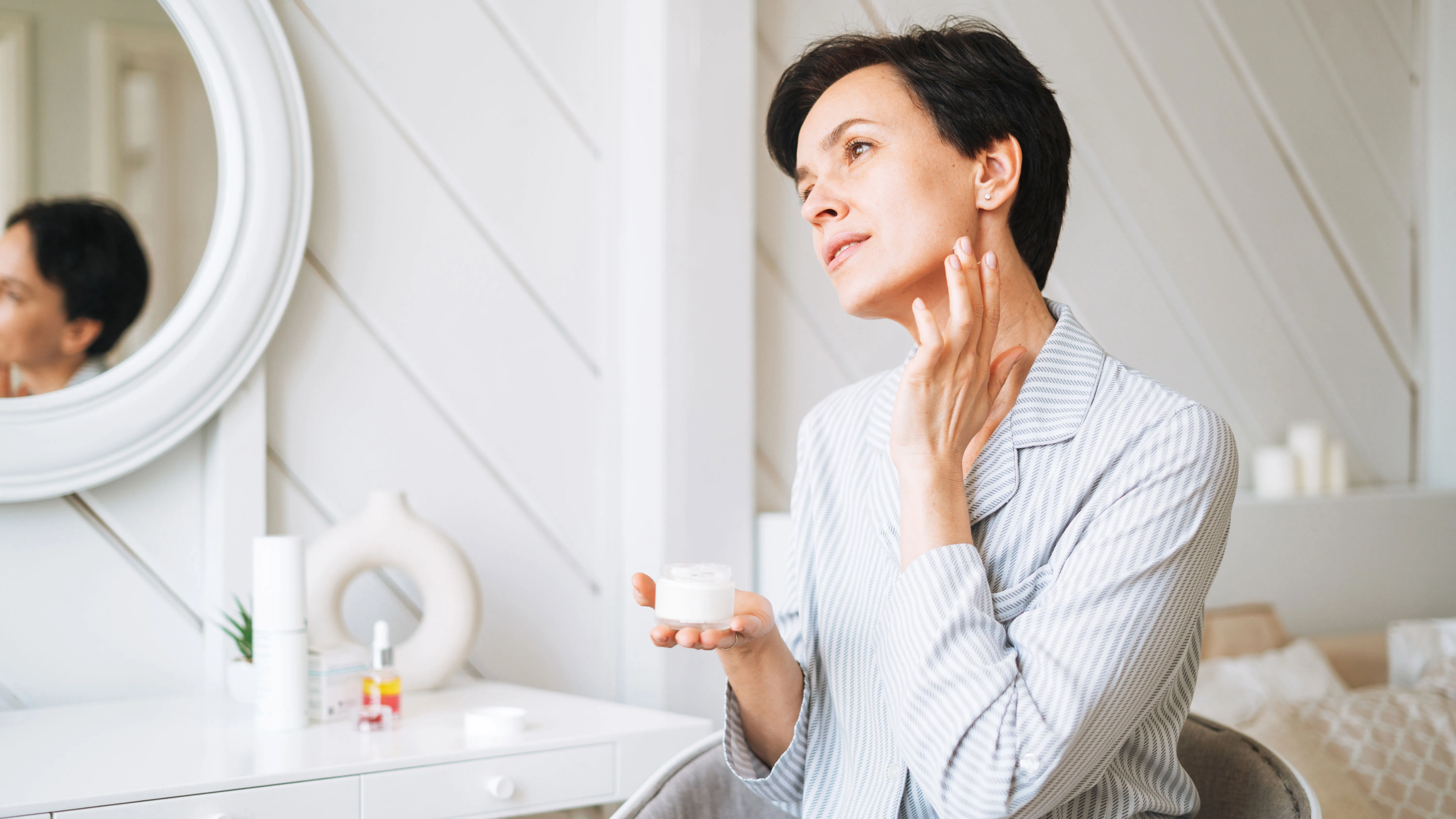 Young adult brunette woman with short hair applies cream to her face at home, morning skincare routine - stock photo