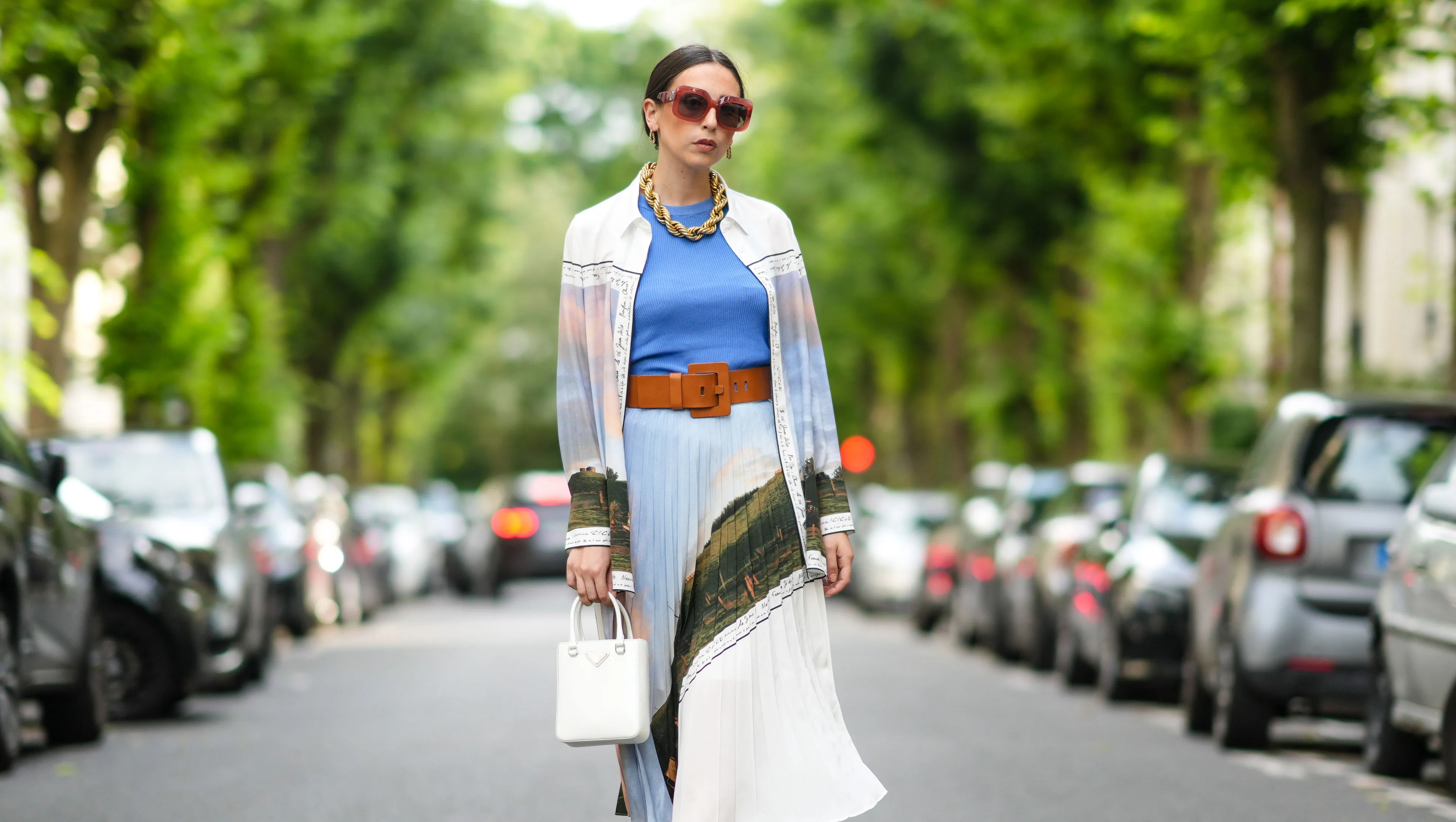 PARIS, FRANCE - AUGUST 02: Gabriella Berdugo wears Loewe sunglasses, a golden large maxi chain necklace, a blue top, a long shirt with colored printed landscape details from Icicle, a matching pleated midi skirt with printed landscape details from Icicle, a white Prada bag, a leather brown large belt from Barbara Bui, green square sandals from Reike Nen, on August 02, 2021 in Paris, France. (Photo by Edward Berthelot/Getty Images)