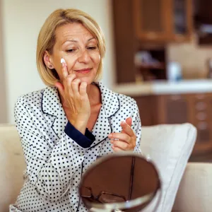 Smiling mature woman applying face cream under her eye while doing skin care routine at home - stock photo