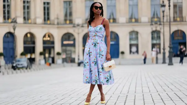 PARIS, FRANCE - APRIL 29: Emilie Joseph wears black large sunglasses, crystal earrings, a pale blue with red / green / navy blue / beige flower print pattern heart neck / tank-top / belted / midi dress, gold bracelets, a beige wickers and white shiny leather bi-material handbag, yellow shiny leather pointed pumps heels shoes , during a street style fashion photo session, on April 29, 2023 in Paris, France. (Photo by Edward Berthelot/Getty Images)