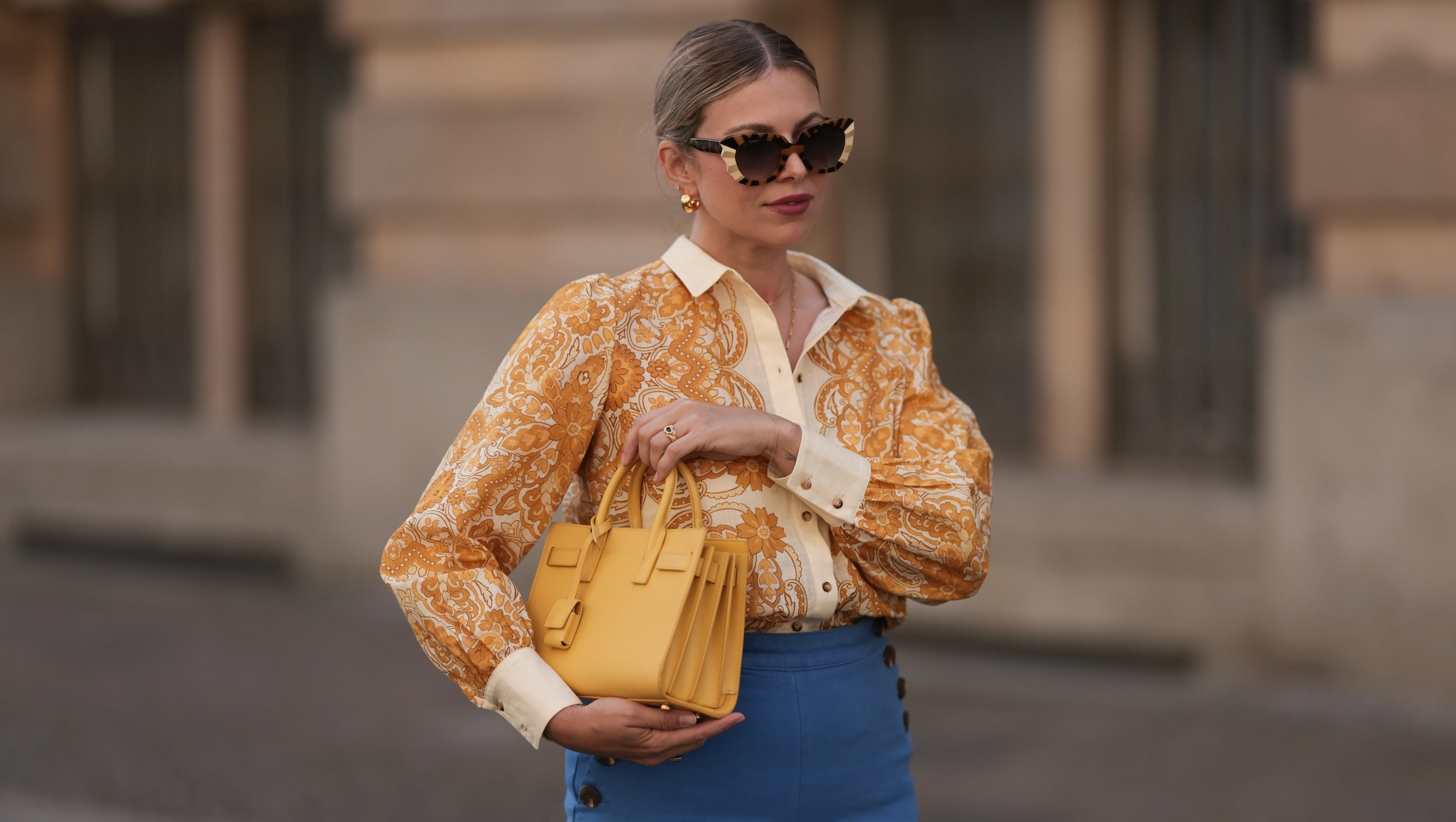 BERLIN, GERMANY - JUNE 07: Aline Kaplan is seen wearing large sunglasses with beige, brown and black pattern from Tibi; chunky golden hoop earrings; several golden necklaces; a creme-white blouse with floral orange pattern from S√©zane; a blue high-waisted pair of trousers with two button borders on the hips from S√©zane; golden rings and a yellow leather handbag from Saint Laurent on June 07, 2024 in Berlin, Germany. (Photo by Jeremy Moeller/Getty Images)