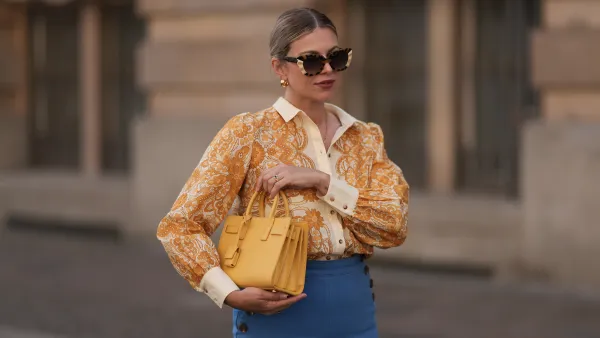 BERLIN, GERMANY - JUNE 07: Aline Kaplan is seen wearing large sunglasses with beige, brown and black pattern from Tibi; chunky golden hoop earrings; several golden necklaces; a creme-white blouse with floral orange pattern from Sézane; a blue high-waisted pair of trousers with two button borders on the hips from Sézane; golden rings and a yellow leather handbag from Saint Laurent on June 07, 2024 in Berlin, Germany. (Photo by Jeremy Moeller/Getty Images)
