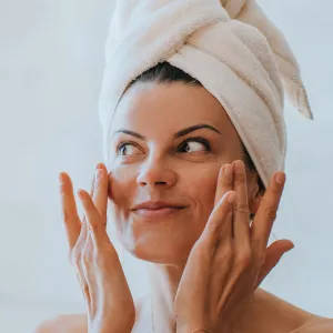 Young adult hispanic woman at bathroom with towel wrapped around head applying cream on face satisfied by spa procedure. Relaxed American female trying new skin care lotion at hotel. Healthy life.