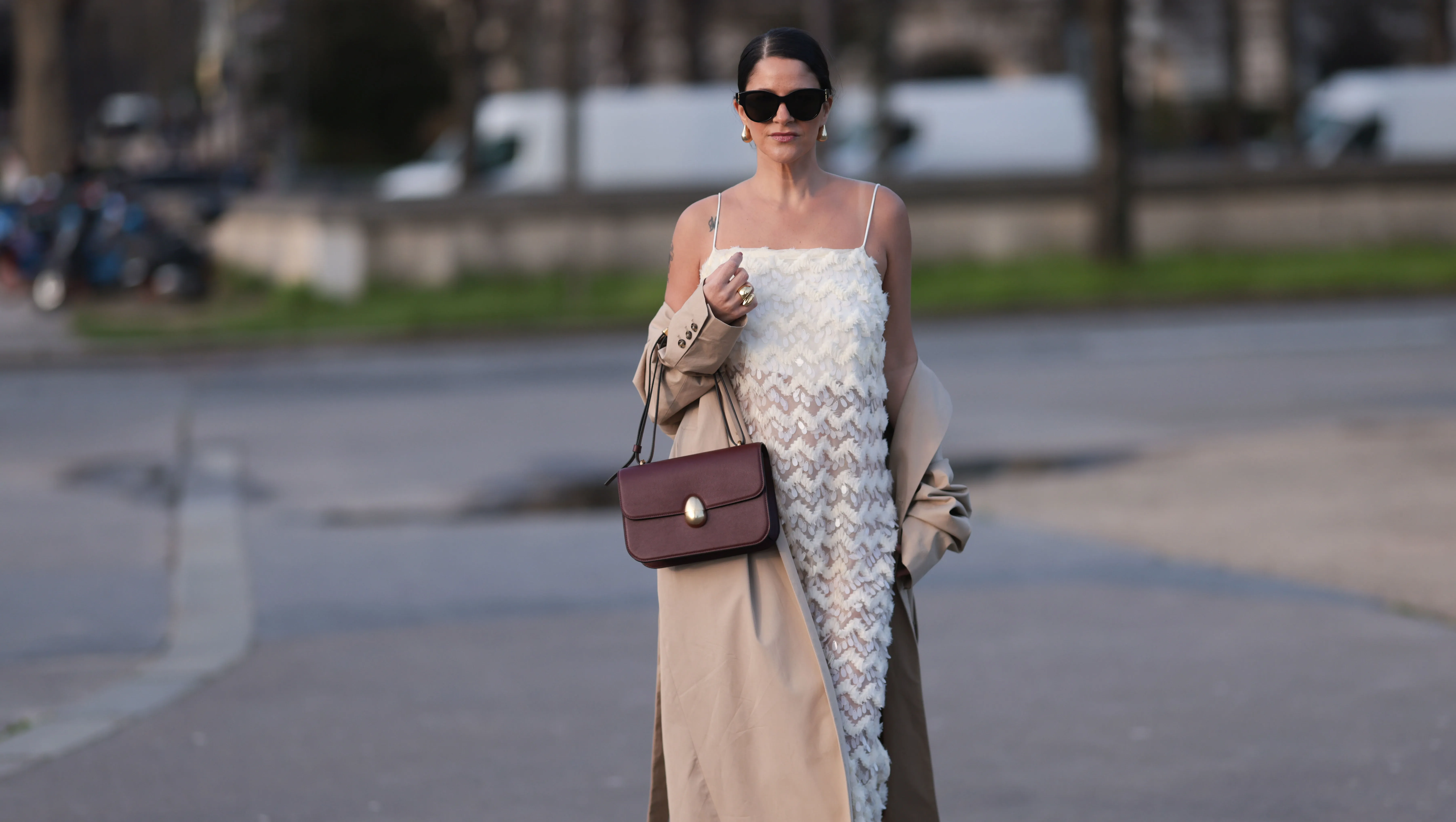 PARIS, FRANCE - FEBRUARY 27: Gili Biegun seen wearing YSL black sunglasses, Bottega Veneta gold tear drops earrings, white long sheer pearl embroidered maxi dress, gold statement rings, beige oversized long coat, Neous burgundy leather bag and burgundy varnished leather pumps / heels, outside Anrealage, during the Womenswear Fall/Winter 2024/2025 as part of Paris Fashion Week on February 27, 2024 in Paris, France. (Photo by Jeremy Moeller/Getty Images)
