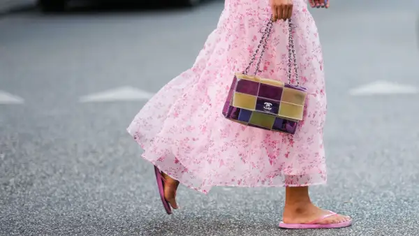 COPENHAGEN, DENMARK - AUGUST 10: A guest wears a white with pale pink flower print pattern / puffy shoulder / short sleeves / ruffled midi dress, a red bracelet, a yellow and purple shiny plastic checkered print pattern handbag from Chanel, pink flip flop , outside Baum und Pferdgarten, during Copenhagen Fashion Week Spring/Summer 2023, on August 10, 2022 in Copenhagen, Denmark. (Photo by Edward Berthelot/Getty Images)