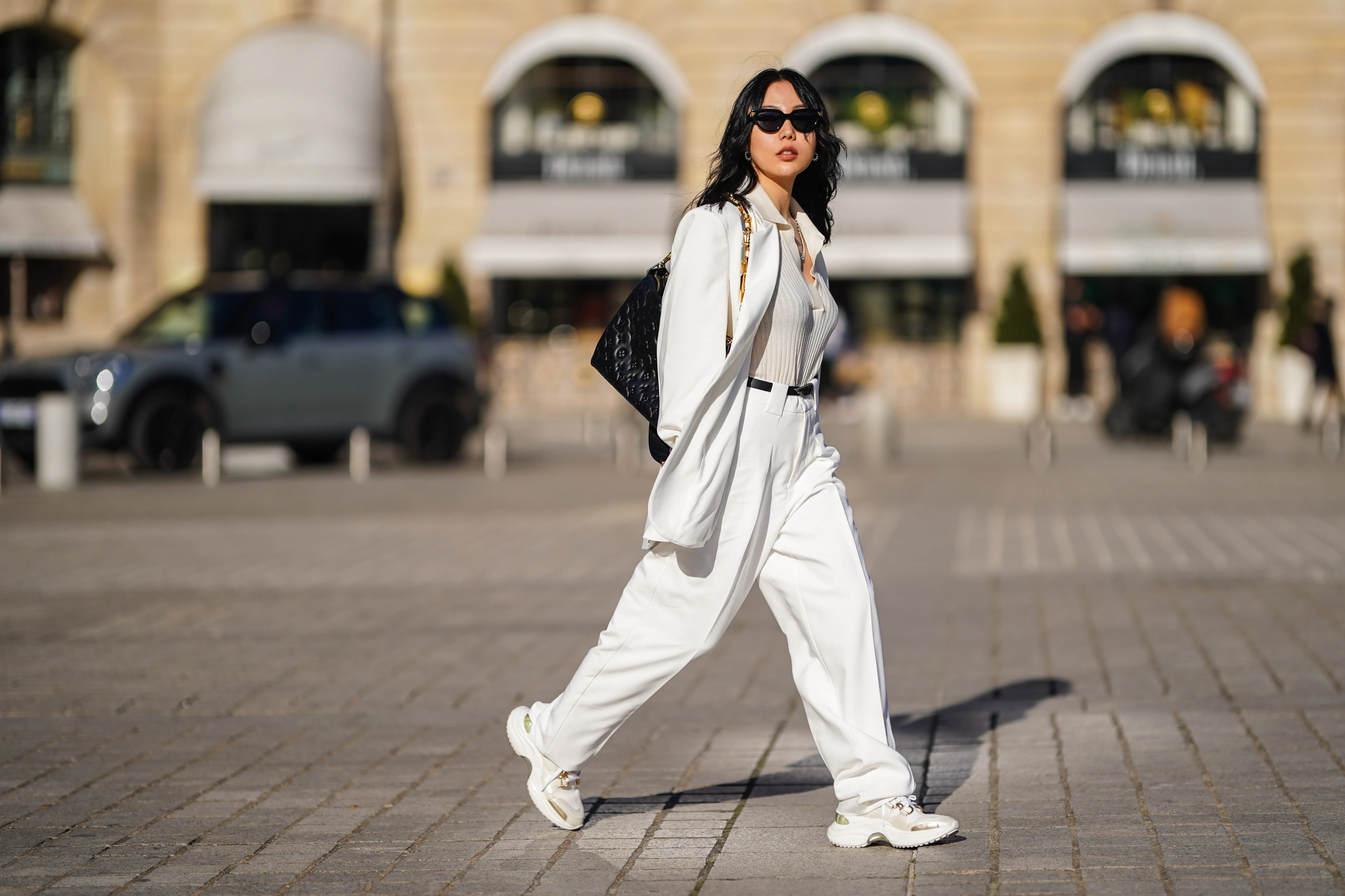 Xiayan wears black Bottega Veneta sunglasses, silver earrings, a silver chain necklace, a white V-neck silk gown, a white long oversized Sundarbay blazer jacket with white suit Sundarbay matching pants, a black shiny leather belt, white and beige with gold details Stellaluna sneakers, a black shiny leather Louis Vuitton monogram-embossed Coussin PM crossbody bag, on February 27, 2021 in Paris, France.