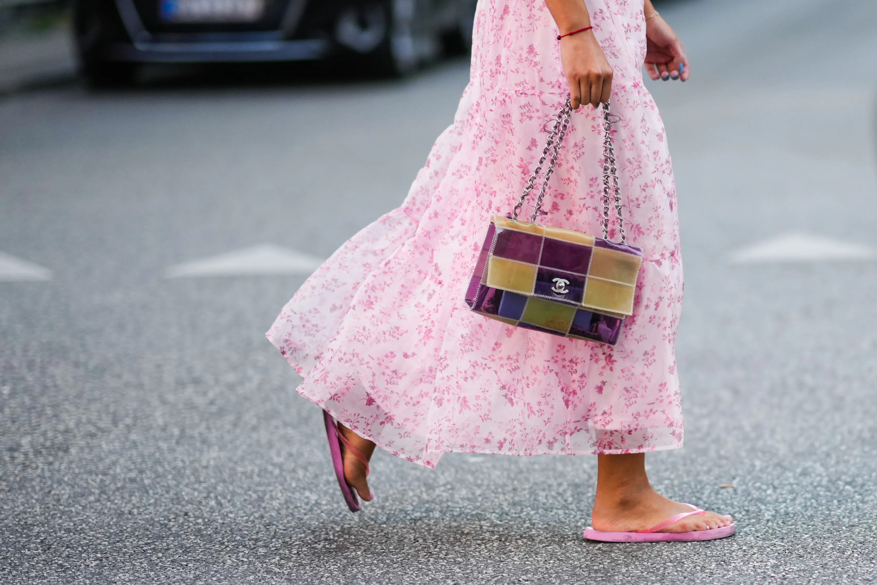 A guest wears a white with pale pink flower print pattern / puffy shoulder / short sleeves / ruffled midi dress, a red bracelet, a yellow and purple shiny plastic checkered print pattern handbag from Chanel, pink flip flop , outside Baum und Pferdgarten, during Copenhagen Fashion Week Spring/Summer 2023, on August 10, 2022 in Copenhagen, Denmark.