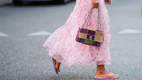A guest wears a white with pale pink flower print pattern / puffy shoulder / short sleeves / ruffled midi dress, a red bracelet, a yellow and purple shiny plastic checkered print pattern handbag from Chanel, pink flip flop , outside Baum und Pferdgarten, during Copenhagen Fashion Week Spring/Summer 2023, on August 10, 2022 in Copenhagen, Denmark.