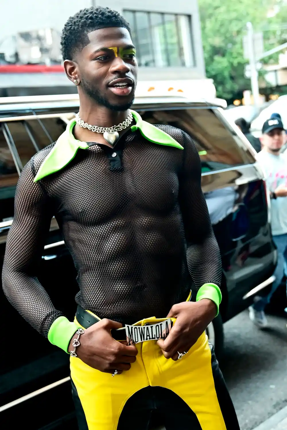Lil Nas X wears black and yellow leather paneled pants and a black mesh polo shirt with brat green elongated lapels