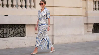 Diane Batoukina wears a white silk midi dress with blue and navy graphic stripe print, short sleeves, and a pointed collar by Sandro. A matching Sandro silk scarf is tied loosely around the neckline. A white mini flap bag with a chain strap and gold hardware by Chanel is worn crossbody. Footwear consists of white open-toe heeled sandals by Aeyde. Dior square sunglasses with dark gradient lenses complete the look. Long, straight brown hair is styled with a center part. Lips are subtly tinted and skin appears smooth and even. Full body shot, during a street style fashion photo session, on April 28, 2025 in Paris, France.