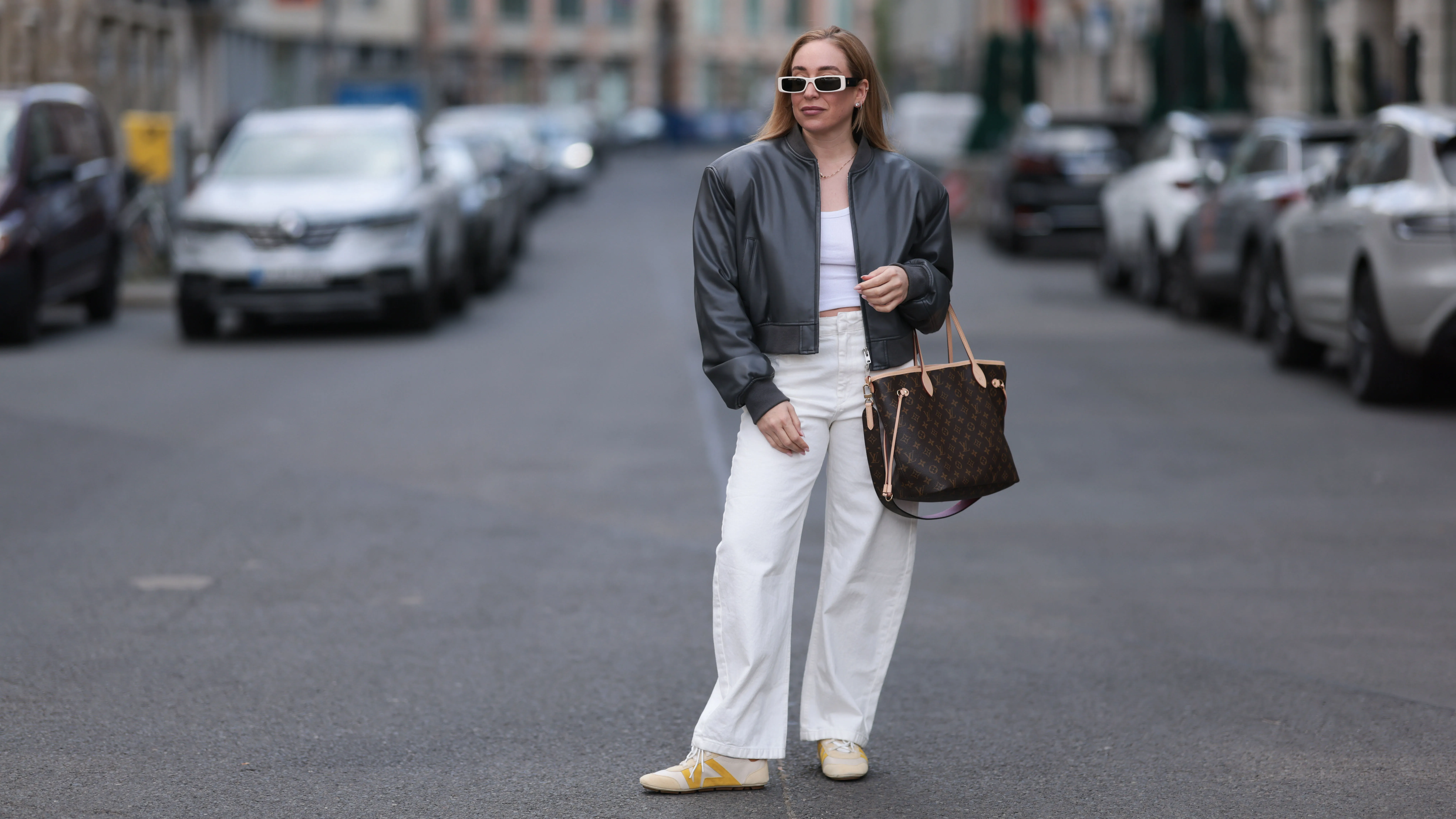 BERLIN, GERMANY - APRIL 09: Sonia Lyson seen wearing Linda Farrow bold white rectangular sunglasses that add a trendy touch, Zara white cotton fitted crop top, Zara high-waisted, wide-leg white relaxed jeans / denim pants, The Frankie Shop black, slightly glossy bomber jacket, Louis Vuitton brown Neverfull tote bag in the classic monogram print and Louis Vuitton beige Sneakerina sneakers / shoes with mustard yellow details, introducing a playful, sporty contrast to the otherwise minimalist outfit, on April 09, 2025 in Berlin, Germany. (Photo by Jeremy Moeller/Getty Images)