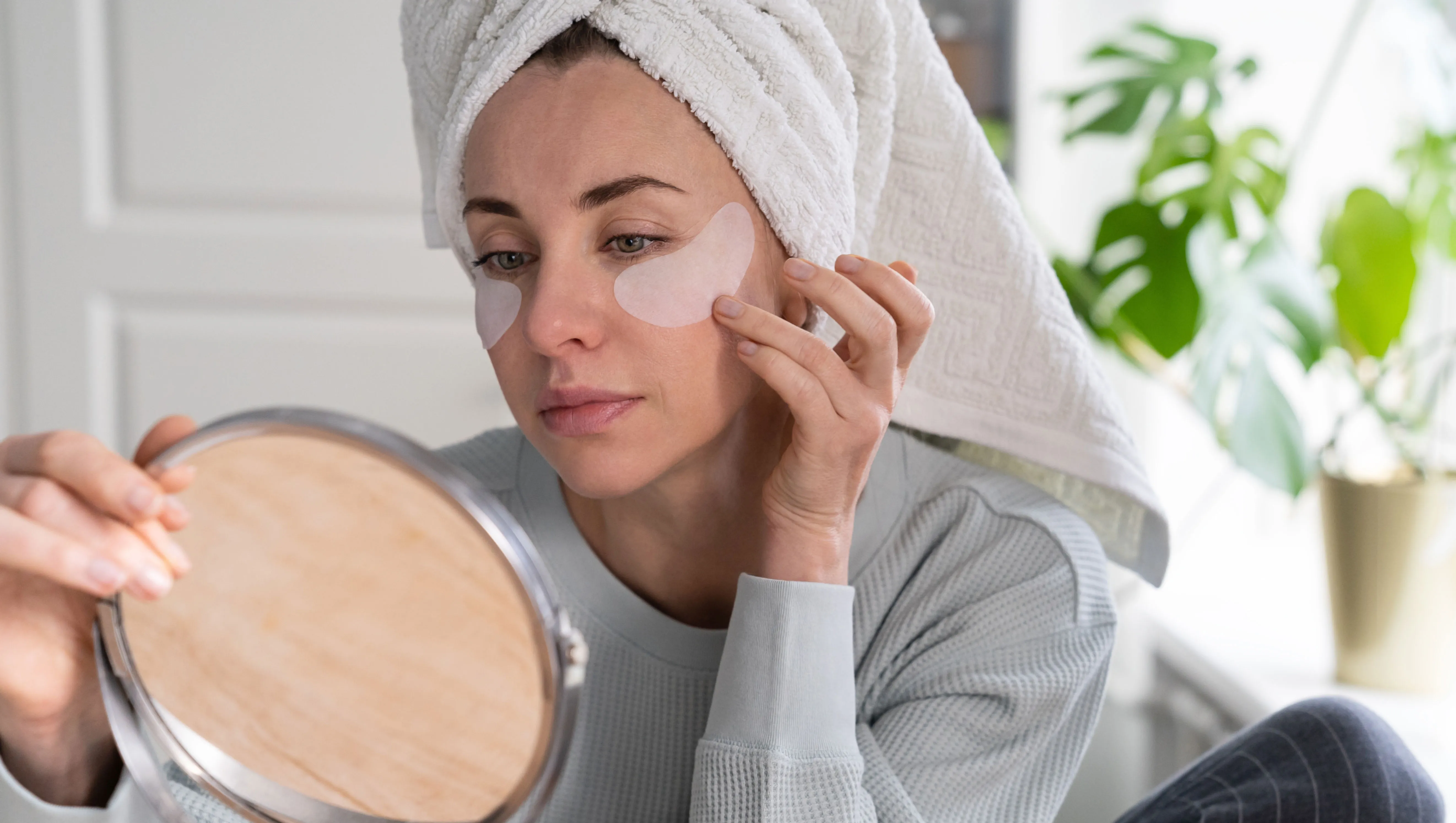 Woman with towel on head applying patches under eyes enriched with collagen, vitamin E, diminishes the signs of aging, helps reduse eye puffiness, looking in mirror. Face skin care beauty at home.
