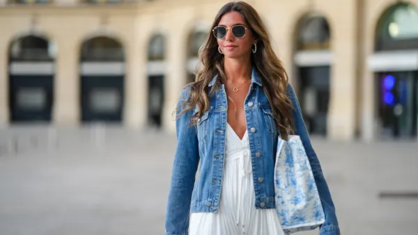 PARIS, FRANCE - AUGUST 20: Amanda Derhy wears earrings, sunglasses, a necklace, a blue denim jacket, a white summer maxi pleated slit off-shoulder dress, during a street style fashion photo session, on August 20, 2022 in Paris, France. (Photo by Edward Berthelot/Getty Images)