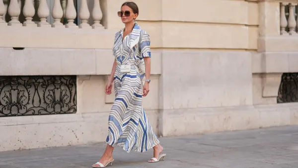 PARIS, FRANCE - APRIL 28: Diane Batoukina wears a white silk midi dress with blue and navy graphic stripe print, short sleeves, and a pointed collar by Sandro. A matching Sandro silk scarf is tied loosely around the neckline. A white mini flap bag with a chain strap and gold hardware by Chanel is worn crossbody. Footwear consists of white open-toe heeled sandals by Aeyde. Dior square sunglasses with dark gradient lenses complete the look. Long, straight brown hair is styled with a center part. Lips are subtly tinted and skin appears smooth and even. Full body shot, during a street style fashion photo session, on April 28, 2025 in Paris, France. (Photo by Edward Berthelot/Getty Images)