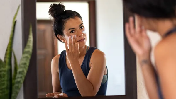 Woman applying white sunblock to face, looking at bathroom mirror - stock photo
