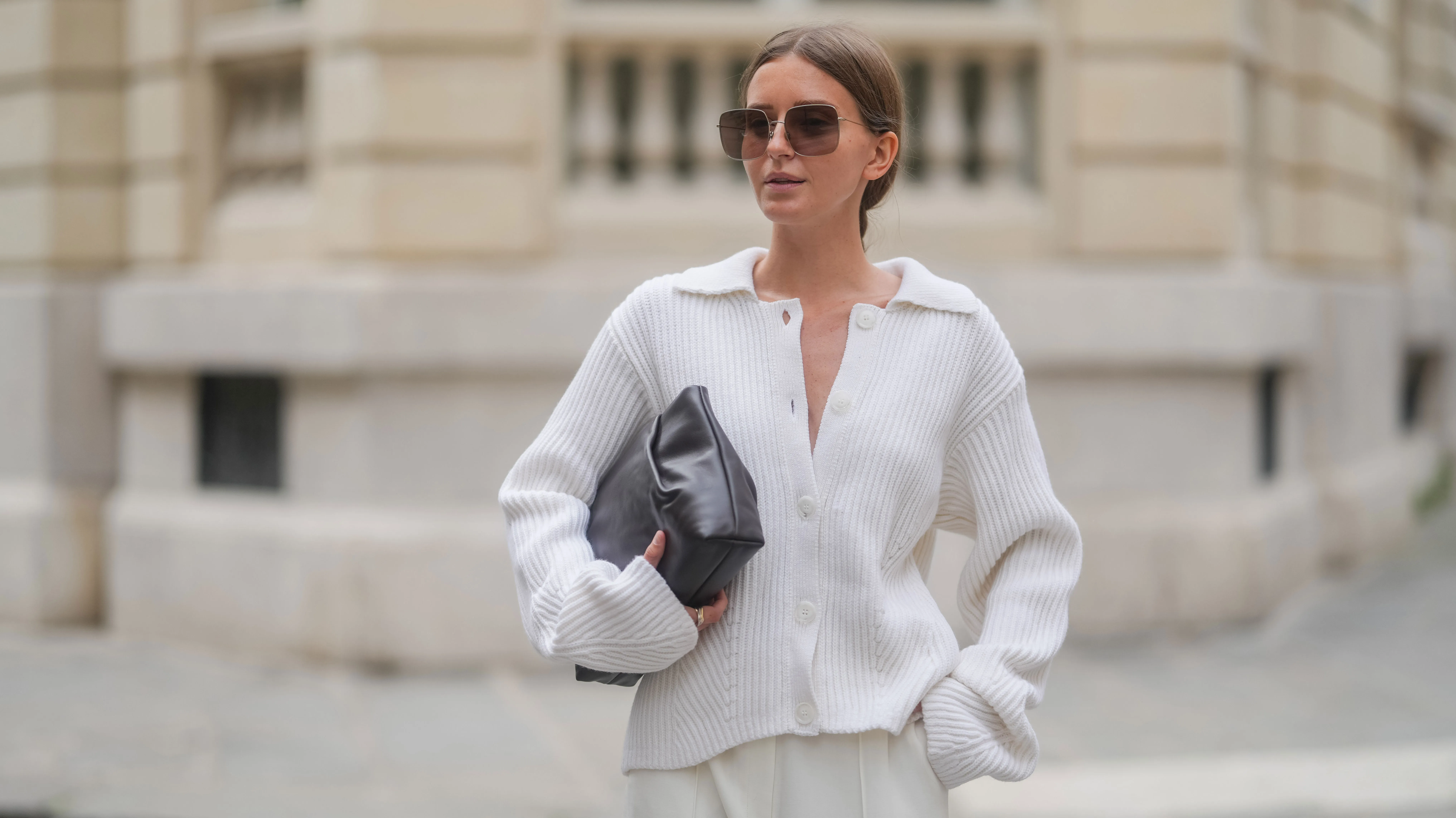PARIS, FRANCE - APRIL 17: Diane Batoukina wears a white ribbed knit cardigan with a wide collar and oversized sleeves by Lovechild 1979, paired with high-waisted ivory trousers by Herskind. A dark brown leather clutch bag by St. Agni is held under one arm. Oversized square Dior sunglasses with brown gradient lenses are worn. Hair is parted at the center and styled in a sleek low bun. Natural makeup with even skin and soft pink lips is visible. Mid shot, during a street style fashion photo session, on April 17, 2025 in Paris, France. (Photo by Edward Berthelot/Getty Images)