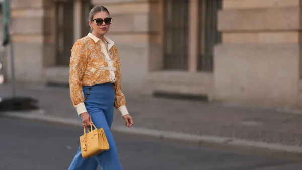 BERLIN, GERMANY - JUNE 07: Aline Kaplan is seen wearing large sunglasses with beige, brown and black pattern from Tibi; chunky golden hoop earrings; several golden necklaces; a creme-white blouse with floral orange pattern from Sézane; a blue high-waisted pair of trousers with two button borders on the hips from Sézane; golden rings; a yellow leather handbag from Saint Laurent and creme-white leather mules from Mango on June 07, 2024 in Berlin, Germany. (Photo by Jeremy Moeller/Getty Images)