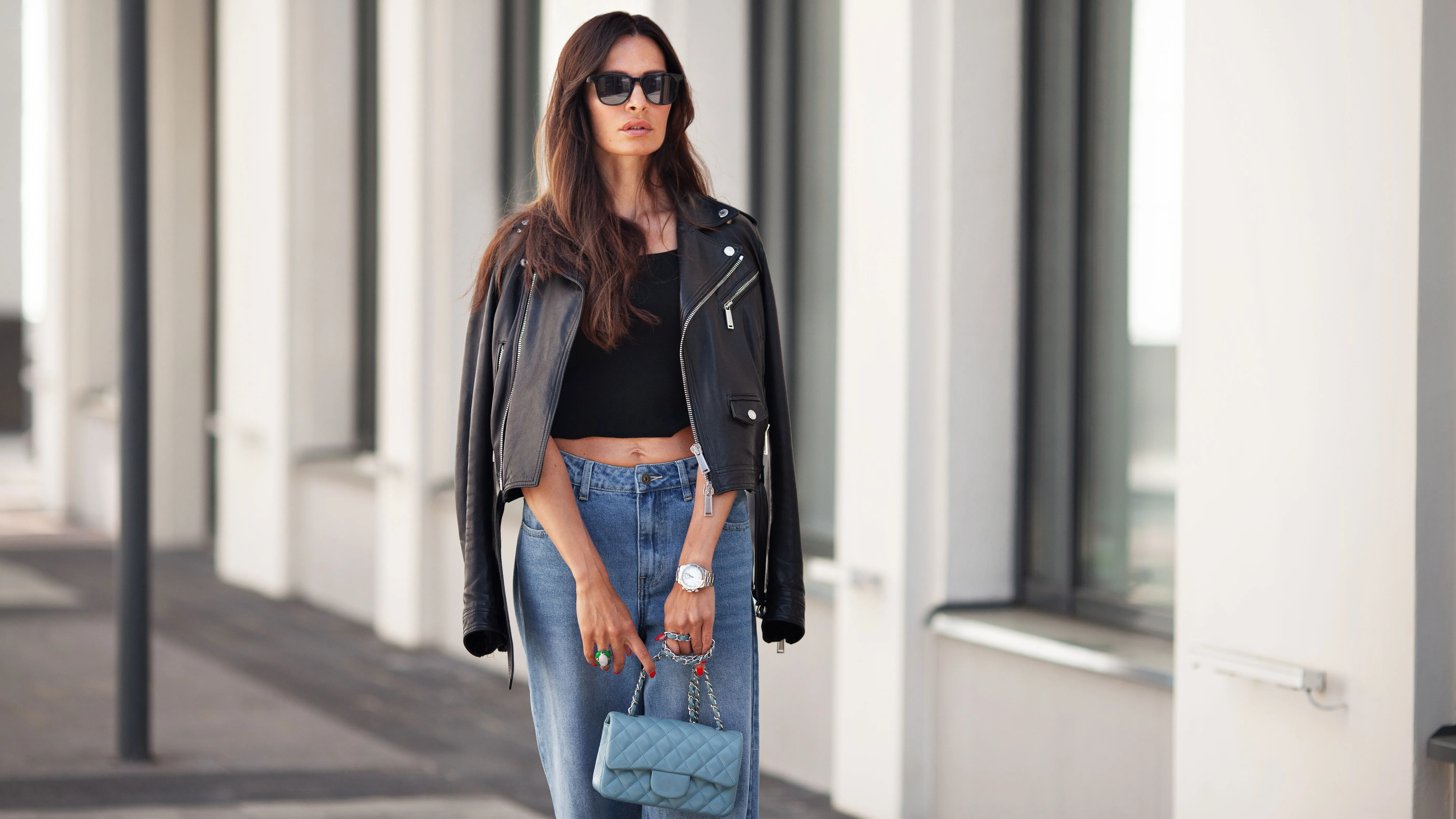 Portrait of gorgeous brunette woman standing city street. Fashion model wears black leather jacket, jeans, short top, small handbag on chain. Street style.