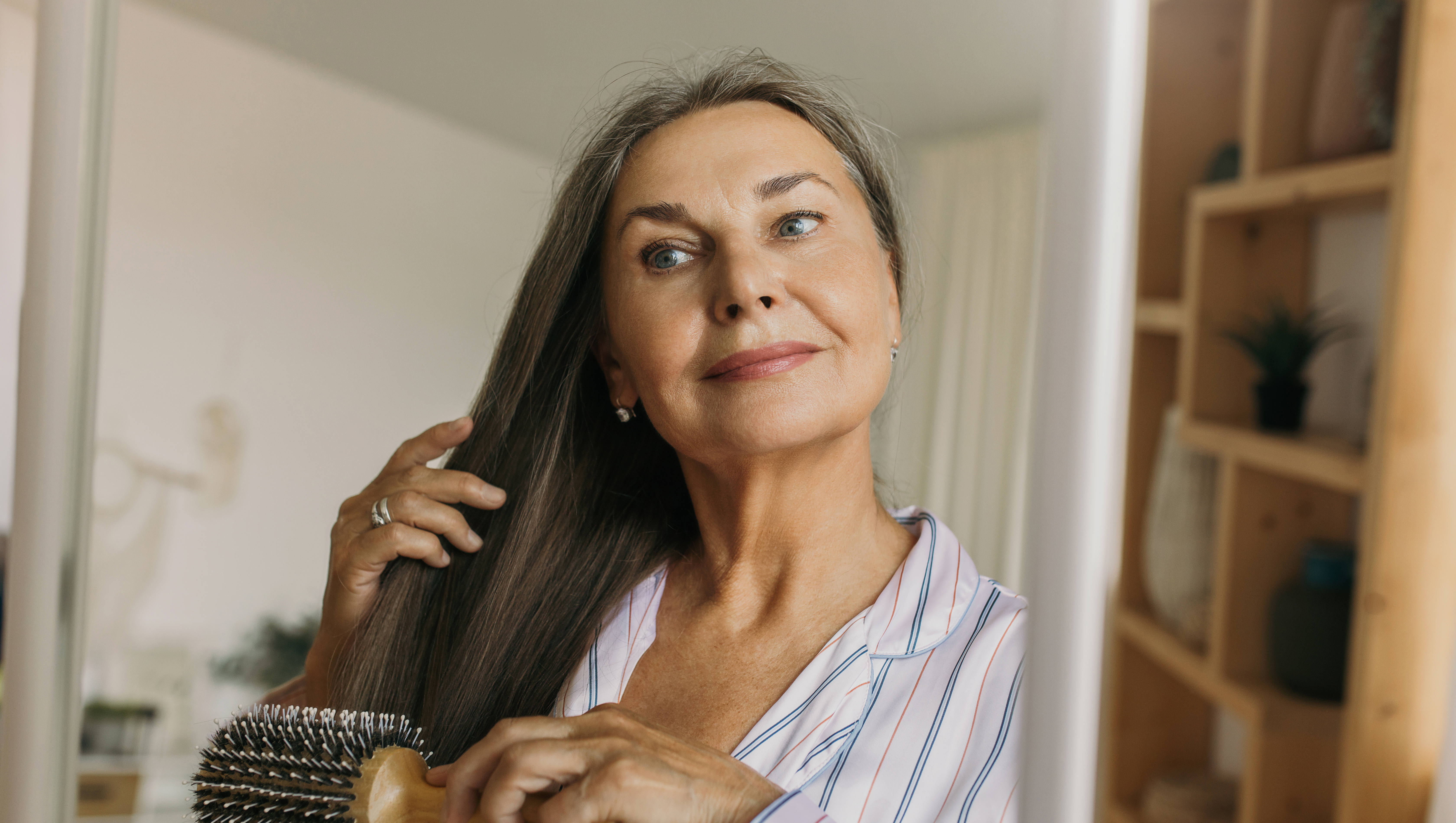 Beautiful, charming elderly female combing grey hair with round hairbrush standing against shelves in hallway or bedroom in front of mirror, looking at her reflection, getting ready to go to bed