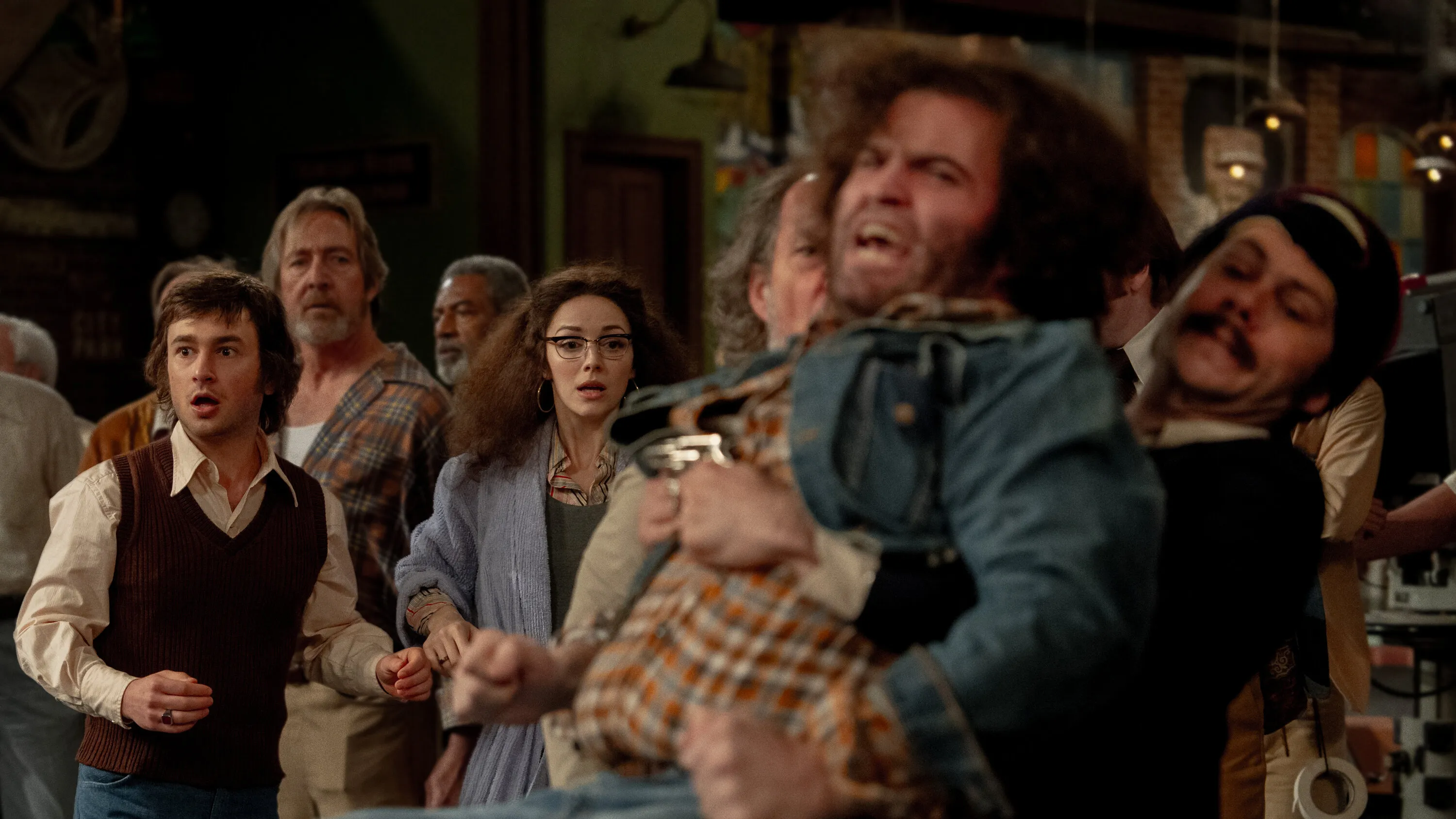 A group of actors look surprised as a man with a mustache holds back an angry man with curly hair in the movie 'Saturday Night.'