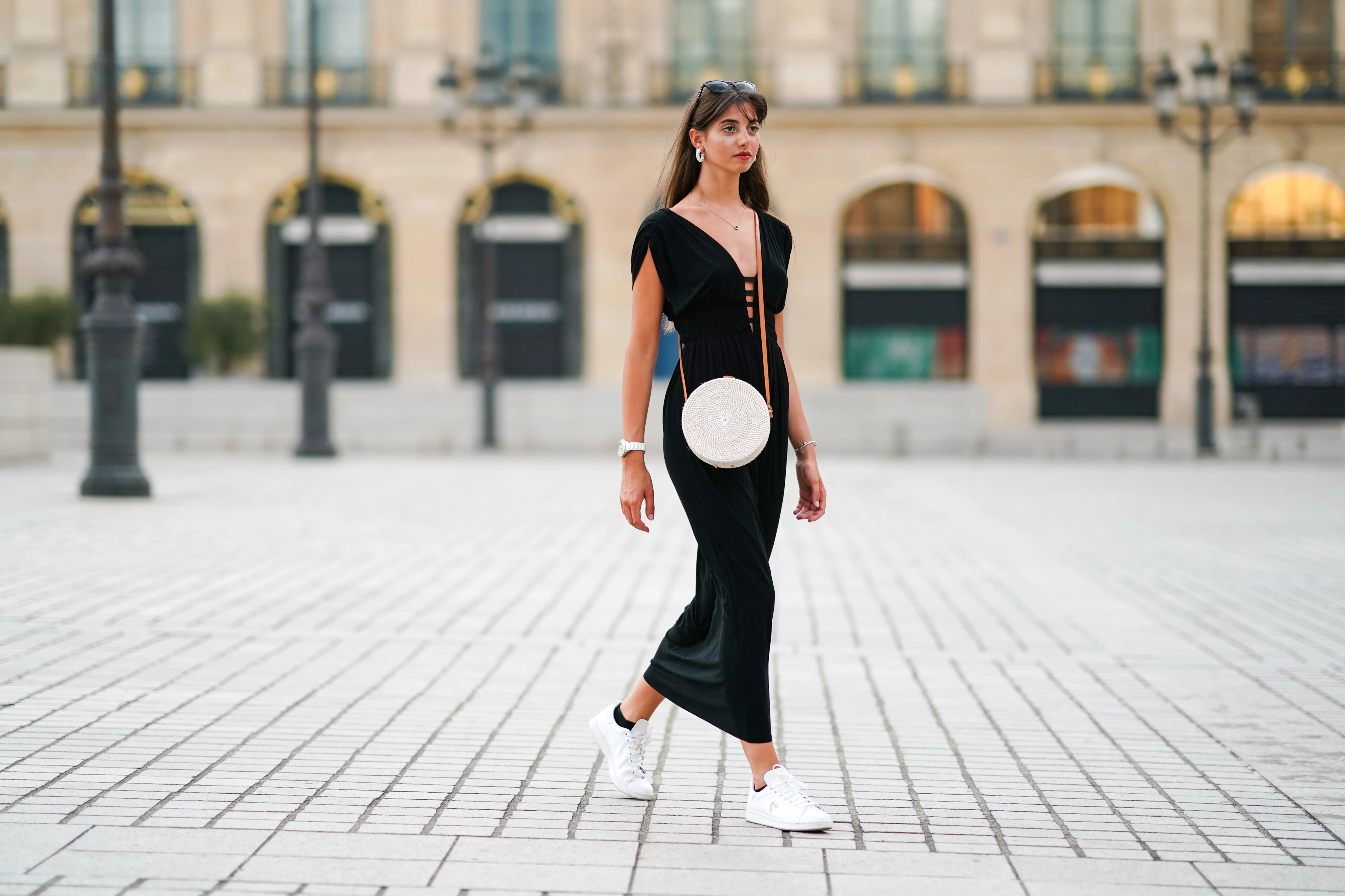 Amanda Derhy wears earrings, a black low-neck v-neck dress, a white circular round straw summer bag, a watch, white Adidas sneakers shoes, on August 09, 2020 in Paris, France.