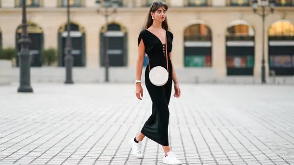 Amanda Derhy wears earrings, a black low-neck v-neck dress, a white circular round straw summer bag, a watch, white Adidas sneakers shoes, on August 09, 2020 in Paris, France.