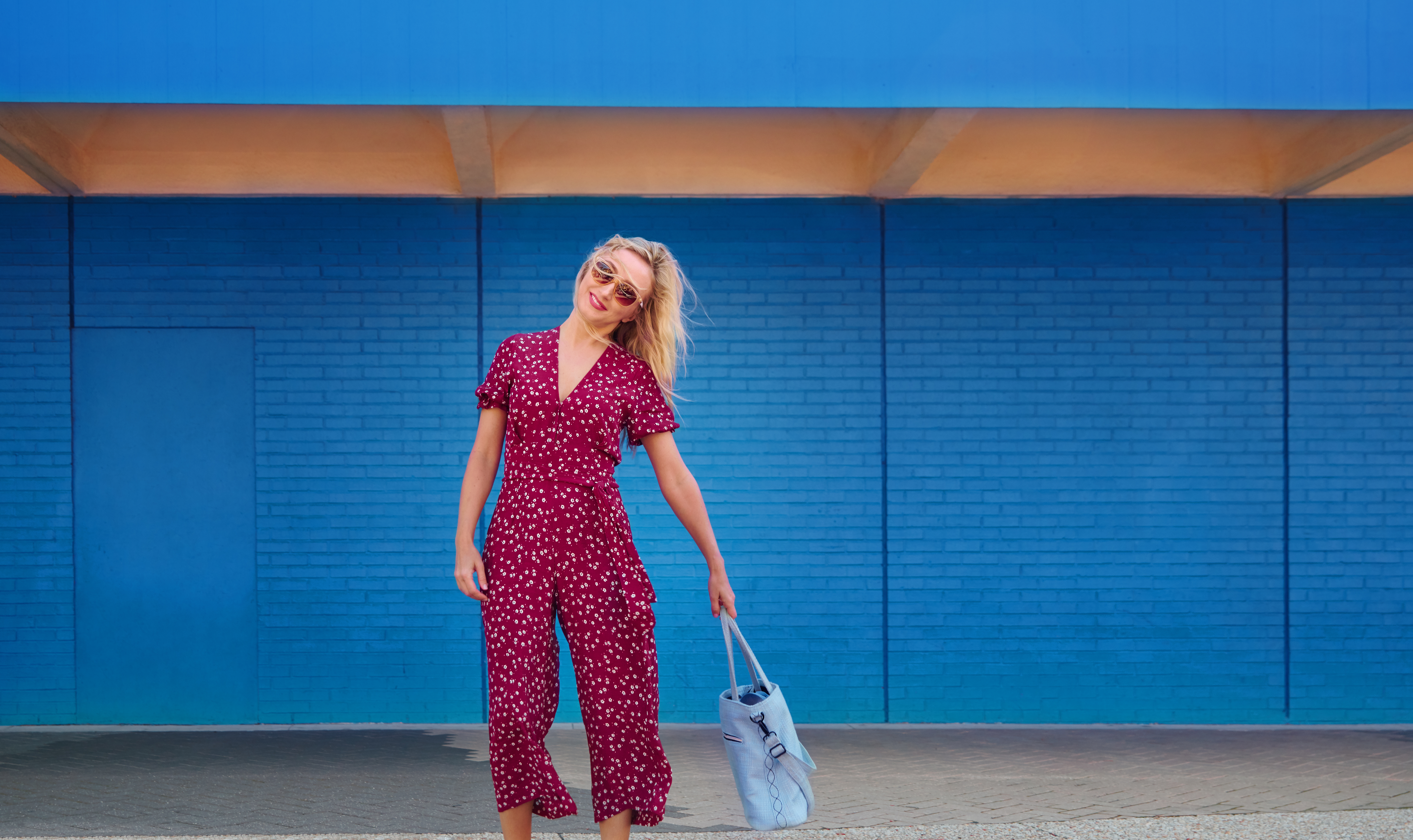 Blond woman wearing burgundy jumpsuit and sunglasses while walking against the blue wall and holding cerulean tote bag