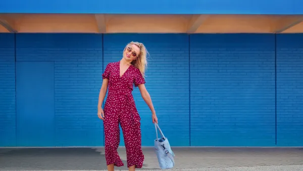 Blond woman wearing burgundy jumpsuit and sunglasses while walking against the blue wall and holding cerulean tote bag