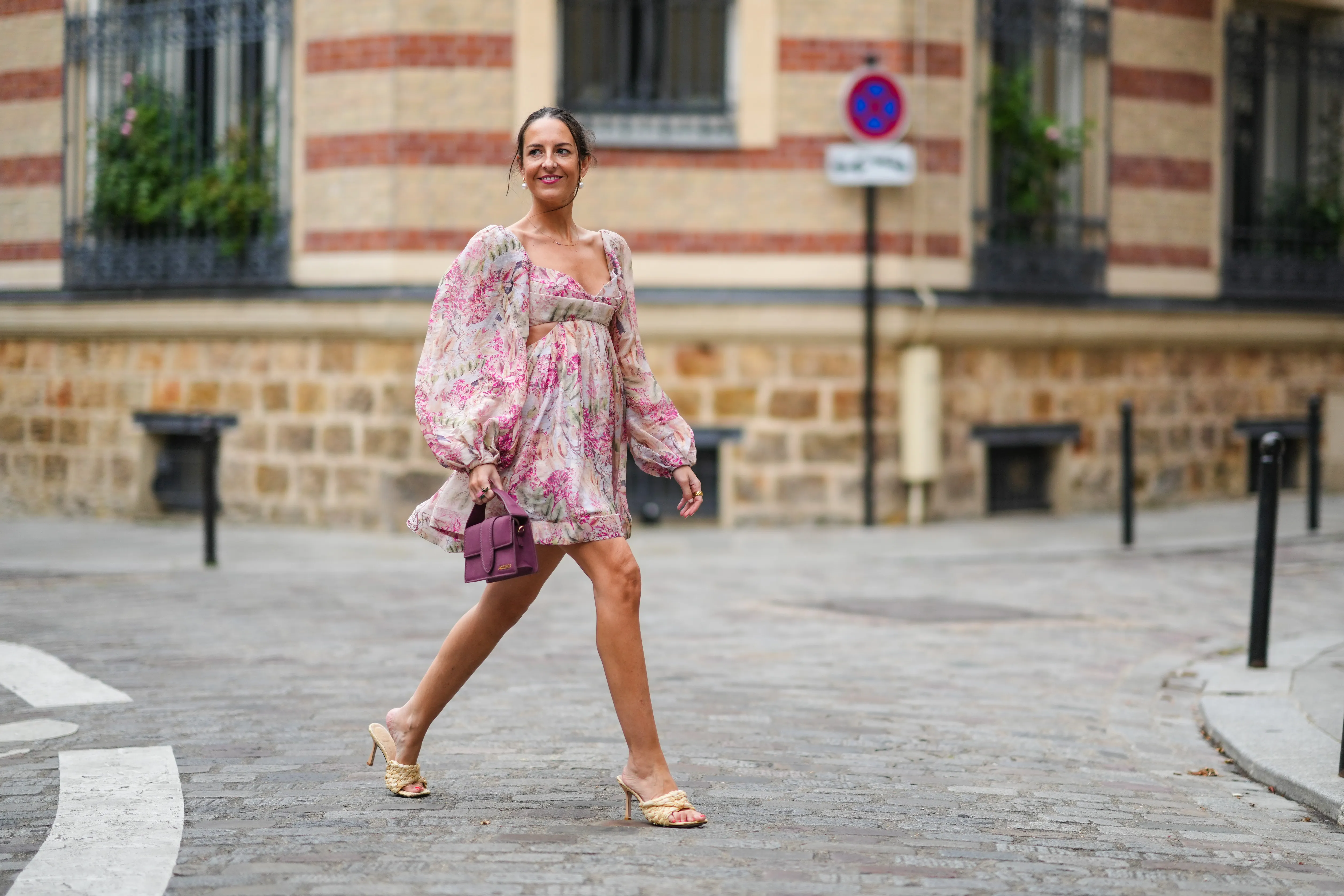Alba Garavito Torre wears earrings, a pale pastel pink mini low-neck gathered and pleated floral print dress with puff shoulders / sleeves and ruffles, a purple Jacquemus bag, Bottega Veneta wicker sandals shoes, on August 04, 2021 in Paris, France.
