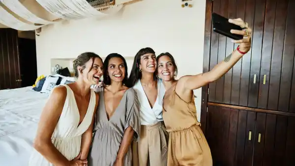 Medium wide shot of smiling and laughing bridesmaids tasking selfie in luxury hotel suite before wedding