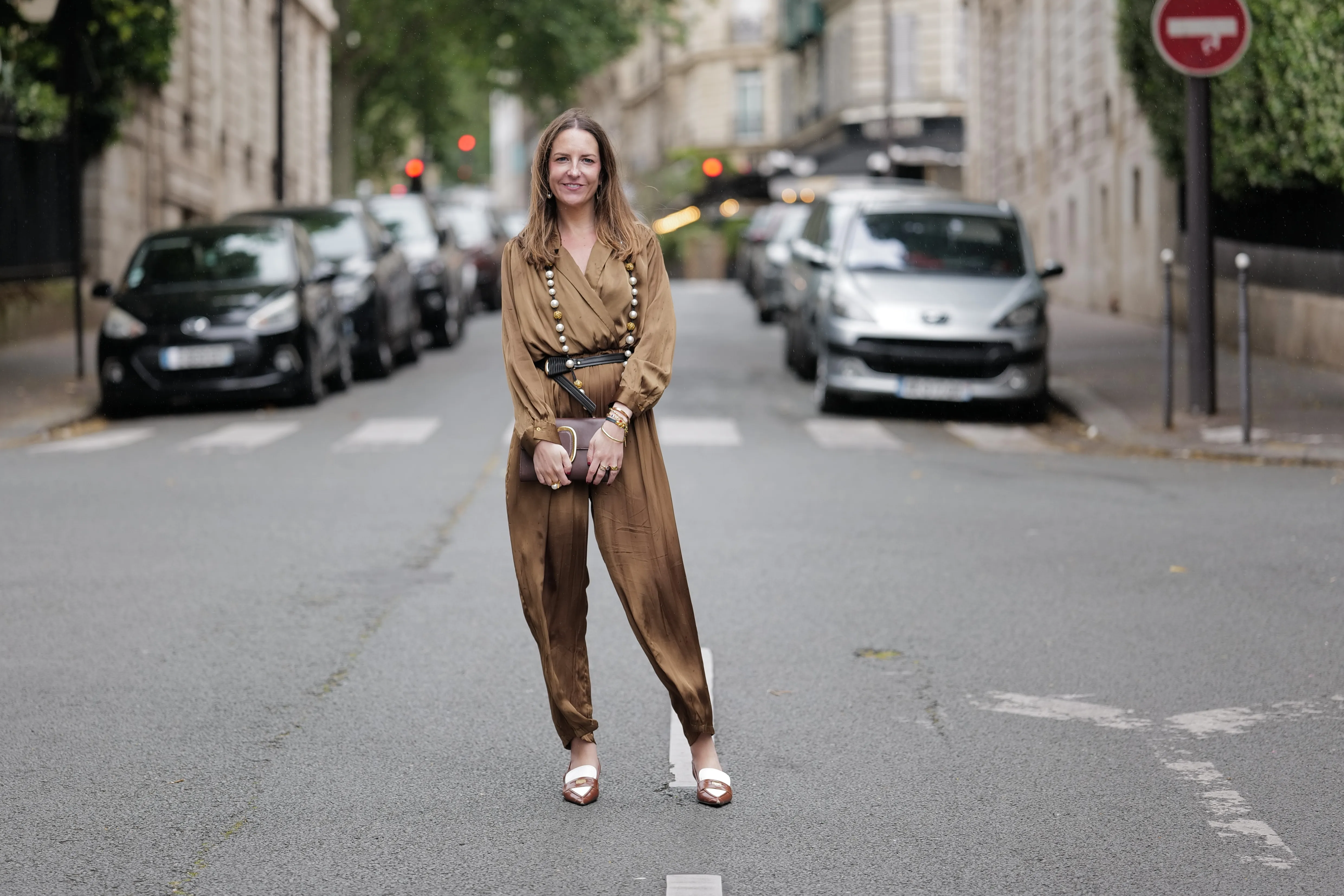 Alba Garavito Torre wears a khaki green long silk jumpsuit from Zara, a brown leather envelope clutch with gold details from Octogony, a brown and white penny loaffers sling backs from Miu Miu, a long pearl and gold necklace from Sempiterno Paris, during a street style fashion photo session, on June 10, 2024 in Paris, France.