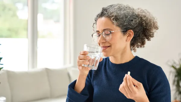 Happy healthy woman of middle age talking pill holding glass of water standing at home. Mature 50 years old lady taking vitamin health care supplement morning daily nutrition treatment concept.